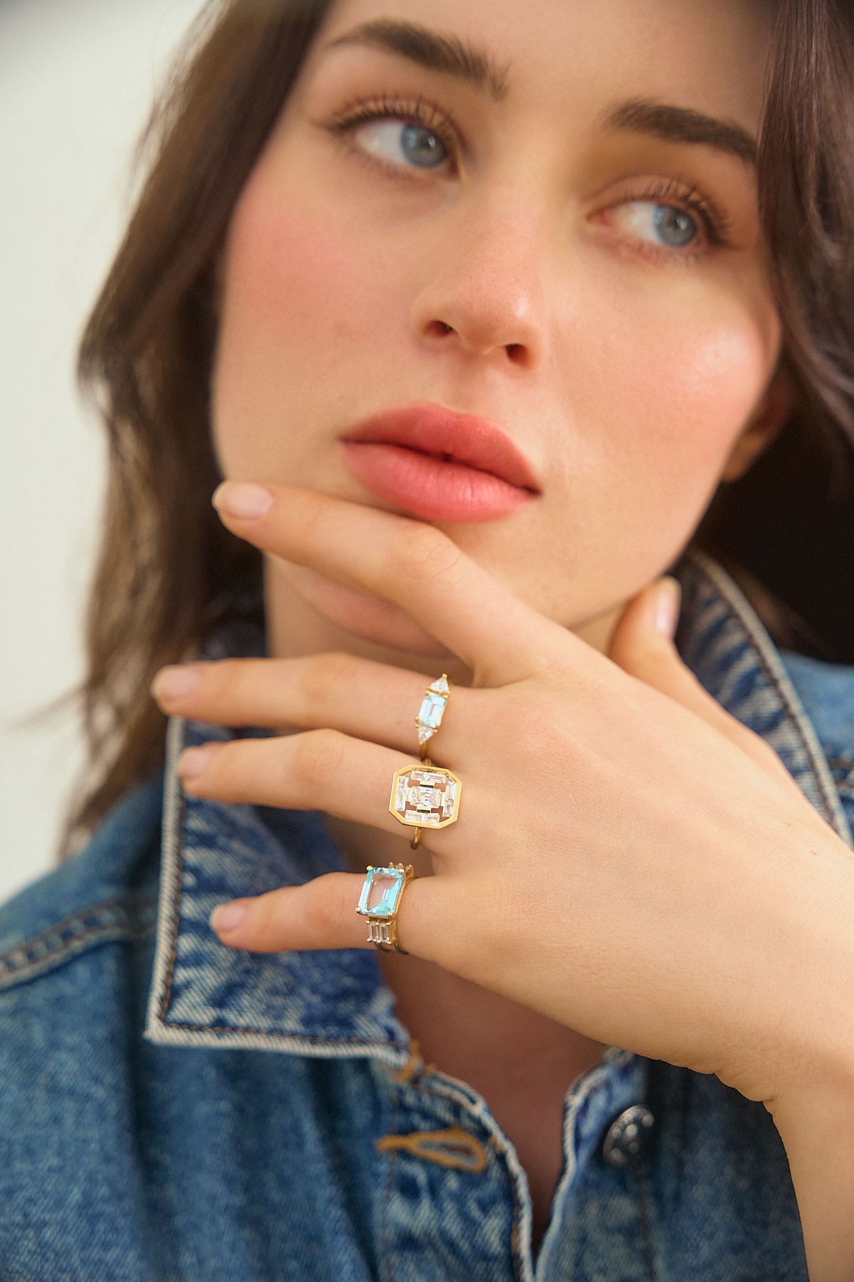 A woman's hand resting on her chin, wearing 3 rings on her fingers from the Art Deco collection by the Nayestones jewelry brand. The rings feature shapes inspired by the Art Deco style reimagined with modernity, showcasing stones such as blue topaz and aquamarine as well as diamonds of various sizes and shapes, in octagon, baguette and triangle.