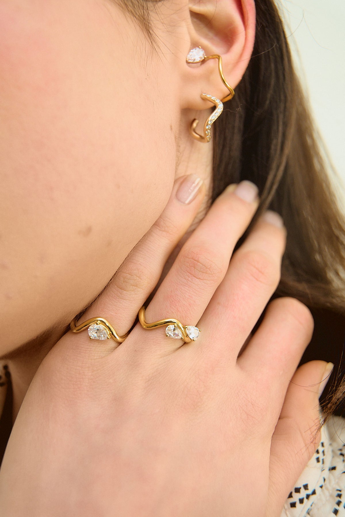 Model's hand resting on her neck in the foreground, wearing two 18K yellow gold rings from the Petite Comète Bold collection by Nayestones, Belgian fine jewelry. The first ring features a central 0.7-carat pear-shaped diamond, and the second two 0.25-carat pear-shaped diamonds facing each other. The model also wears a diamond-paved earring and a gold earcuff adorned with a pear-shaped diamond from the same collection.