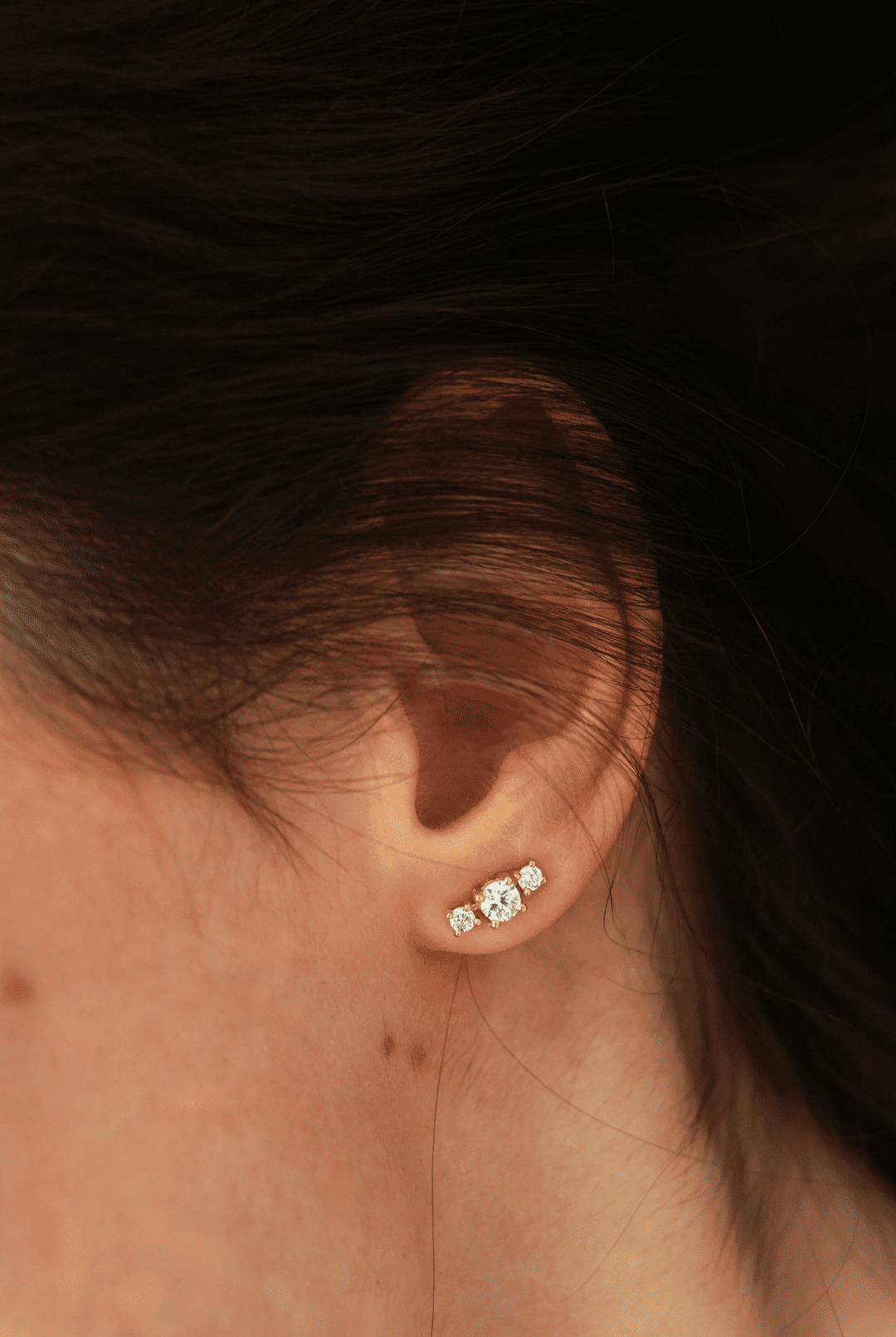 Close-up of an ear featuring a 3-diamond earring, the center one being larger than the two adjoining diamonds. From the Essentials collection by Nayestones, a Belgian fine jewelry brand.