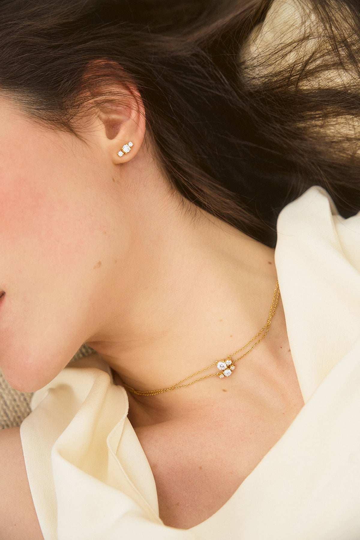 Woman wearing two yellow gold necklaces and earrings set with three diamonds, from the Essential collection by Nayestones. Fine, luminous jewelry worn on bare skin in natural light.