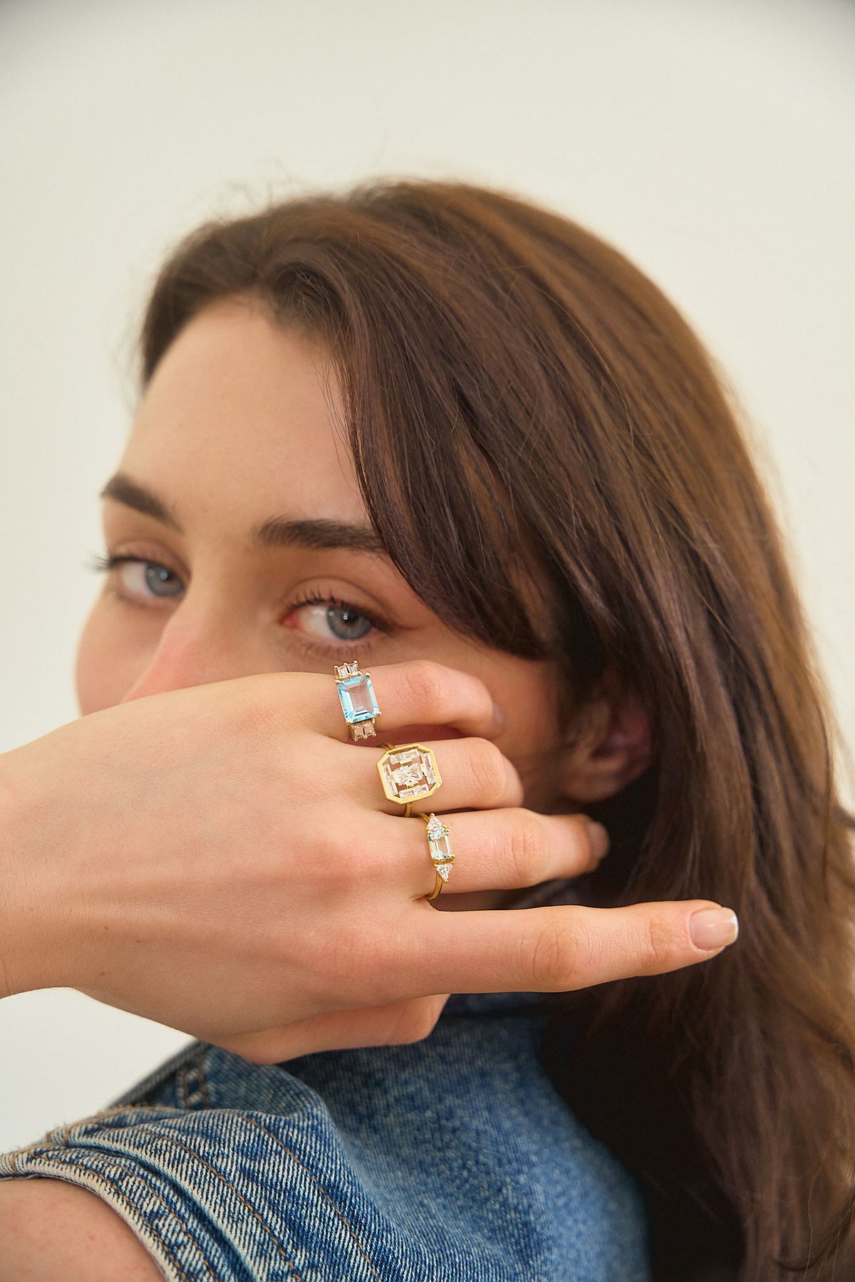 A woman's hand hiding her face, wearing 3 rings on her fingers from the Art Deco collection by the Nayestones jewelry brand. The rings feature shapes inspired by the Art Deco style reimagined with modernity, showcasing stones such as blue topaz and aquamarine as well as diamonds of various sizes and shapes, in octagon, baguette and triangle.