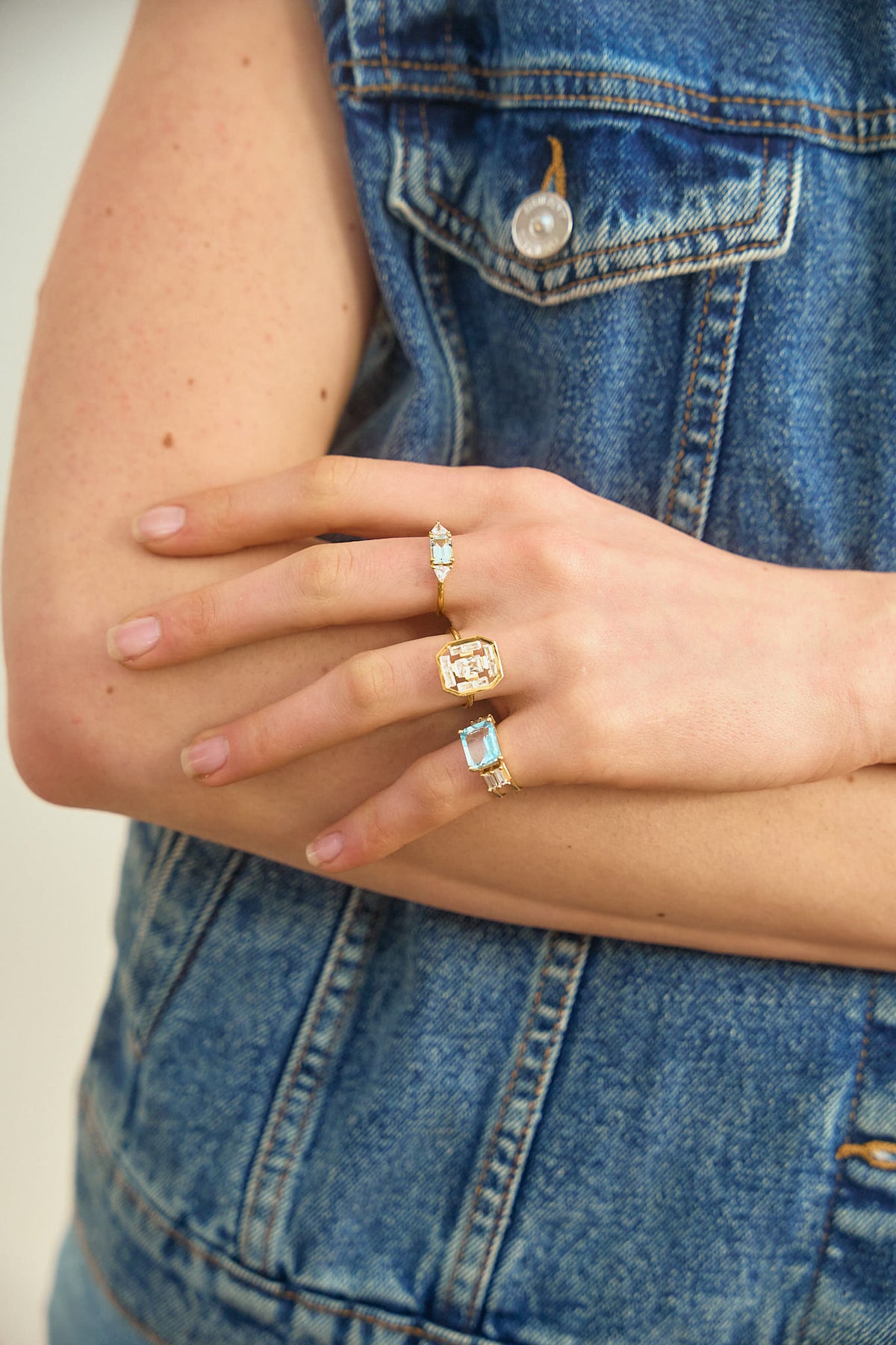 A woman's hand resting on her arm, wearing 3 rings on her fingers from the Art Deco collection by the Nayestones jewelry brand. The rings feature shapes inspired by the Art Deco style reimagined with modernity, showcasing stones such as blue topaz and aquamarine as well as diamonds of various sizes and shapes, in octagon, baguette and triangle.