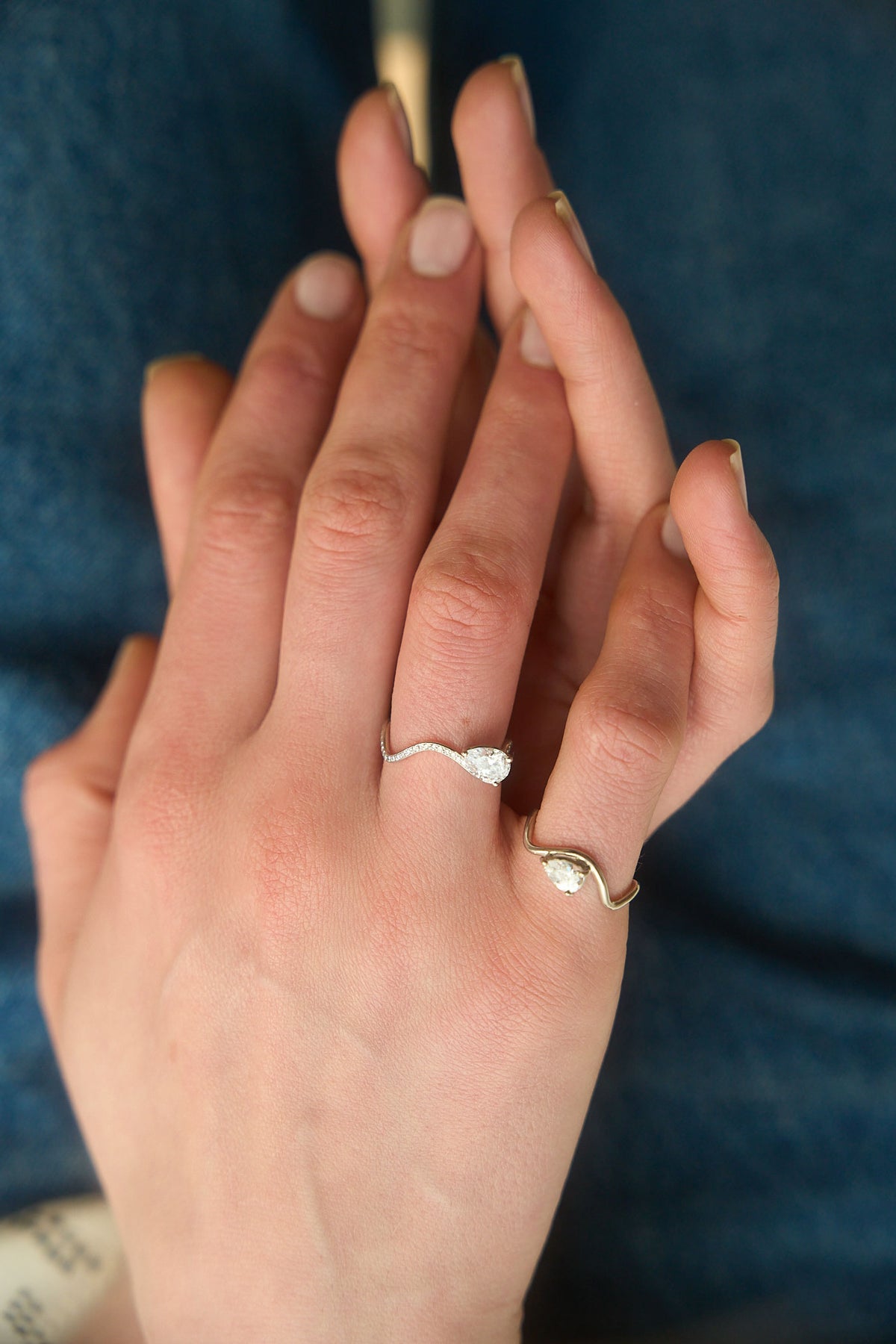 Deux mains se frôlent, l'une portant une bague en or blanc 18 carats avec un diamant central en forme de poire de 0.25 carat à l'auriculaire, et une bague en or blanc pavée de diamants avec un diamant de 0.7 carat à l'annulaire. 