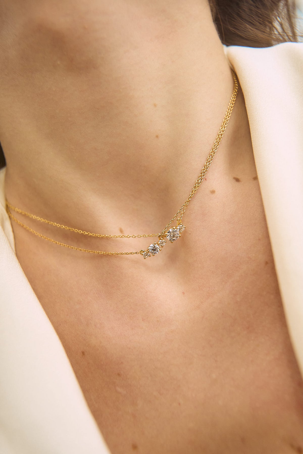Close-up of a neck wearing two overlapping 18K gold necklaces, one with two diamonds and the other with 3 diamonds, the central diamond being larger than the other two. The atmosphere is luminous, reflecting the elegance and minimalism of the Essentials collection by Nayestones, Belgian fine jewelry.