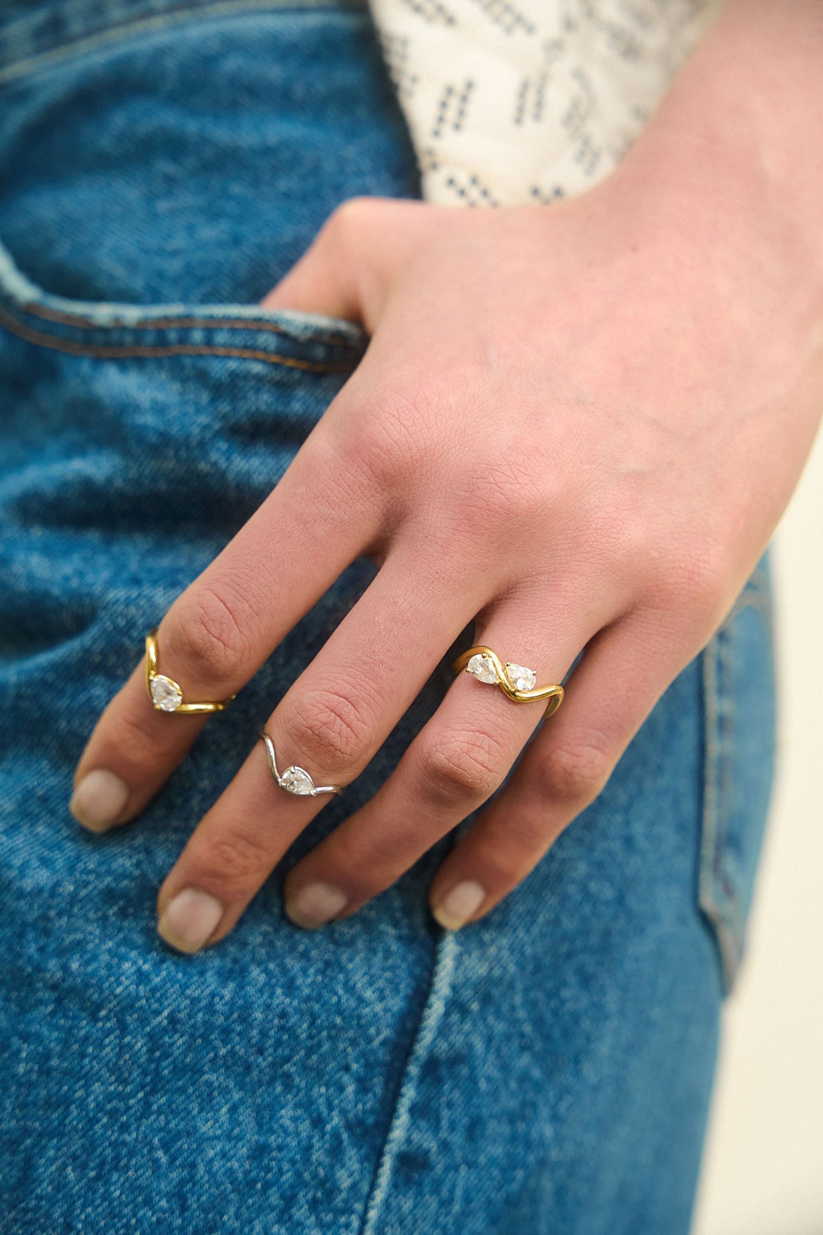 Hand resting on jeans wearing 3 rings from the Petite Comète collection by Nayestones on different fingers. The three rings are in 18K, yellow and white gold, and are set with 0.25 and 0.7 carat pear-shaped diamonds.