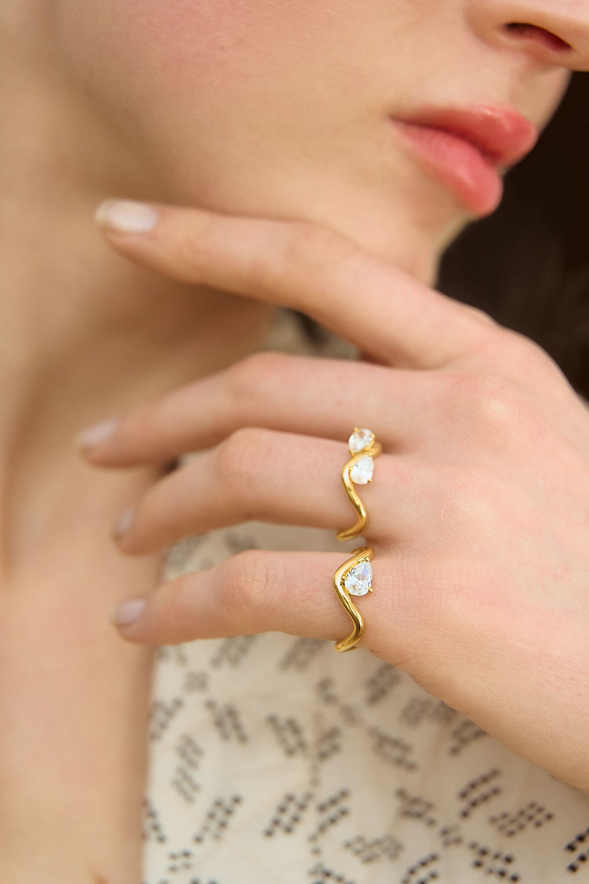 Model's hand resting on her chin, wearing two 18K yellow gold rings from the Petite Comète Bold collection by Nayestones, Belgian fine jewelry. The first ring features a central 0.7-carat pear-shaped diamond, and the second two 0.25-carat pear-shaped diamonds facing each other.