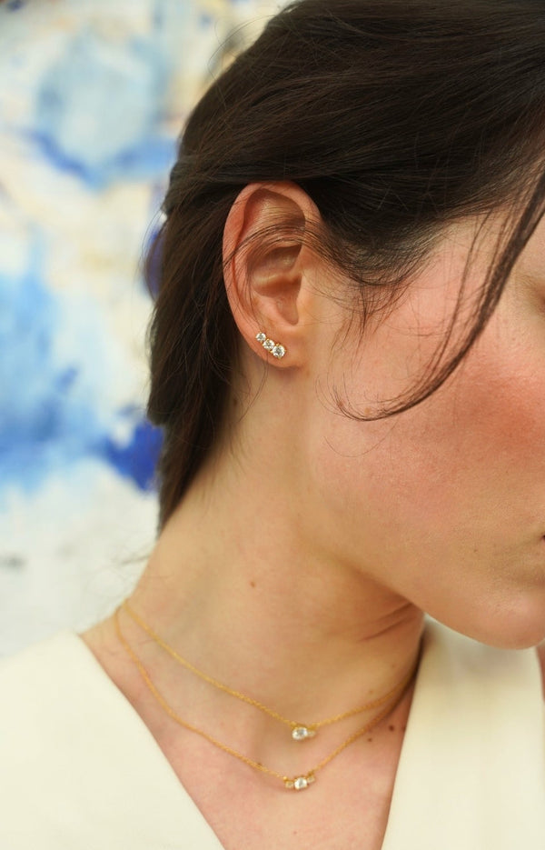 Woman's face in profile wearing an 18K gold earring from the Essential Bridal collection by Nayestones, with 3 diamonds of increasing size, offering timeless elegance and sparkle. Nayestones